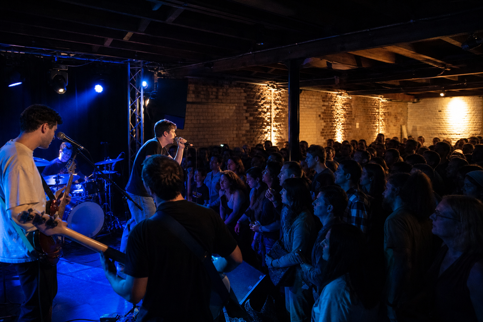 Illustrative independent music room with a low stage, emerging performers, and a close audience under blue light.